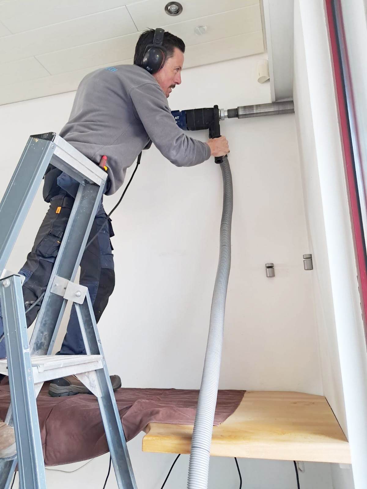 Fabrice staat op een ladder en boort een groot gat in de muur.