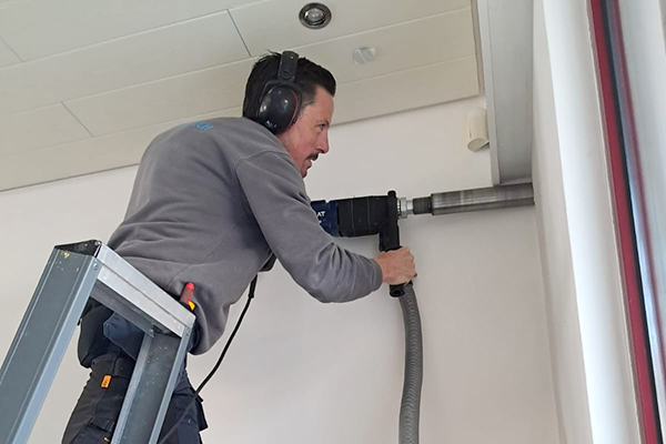 Fabrice maakt op een ladder een groot gat met een boormachine in een muur om de kabels van de airco door te steken.